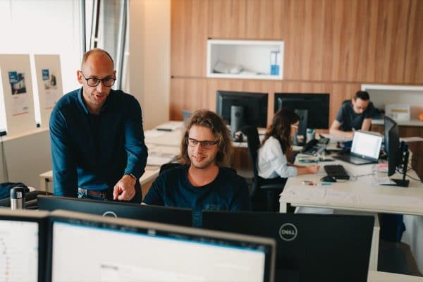 Twee collega's van edibo zijn in overleg met elkaar op hun kantoor. De ene zit achter zijn computer, de andere staat langs hem en kijkt mee op het scherm.